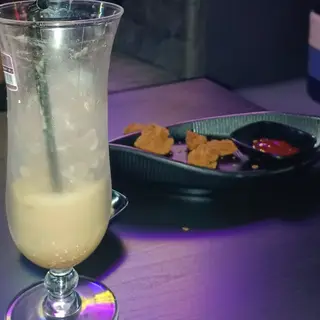 Minuman sejuk berkrim dalam gelas tinggi, dengan sepinggan snek di belakang.