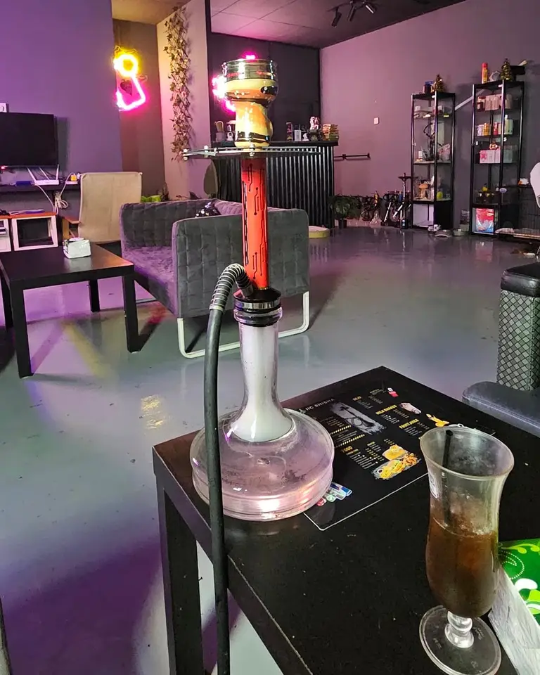 Shisha dan minuman sejuk di atas meja rendah, dengan lampu neon lounge di belakang.